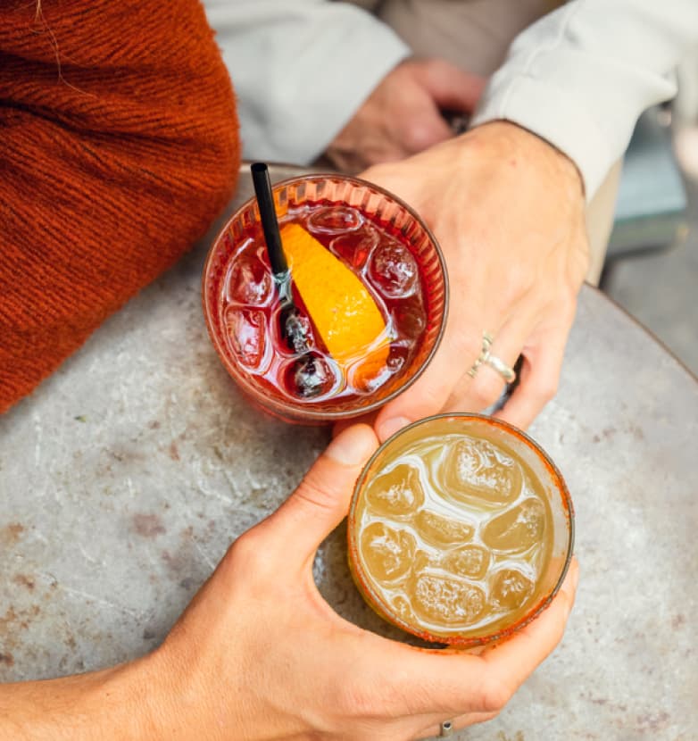 Two people sit at a table in the Wildlight Yulee Florida community, holding cocktails—one a red drink with an orange slice, the other yellowish—their hands close together on a grey surface.