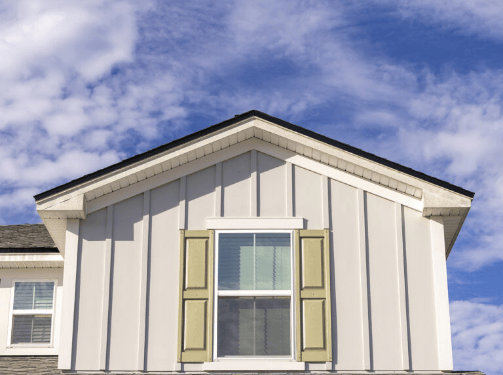 The upper front exterior of a house in the Wildlight Yulee Florida community features vertical siding, a window, pale green shutters, and a gabled roof set against a partly cloudy sky.