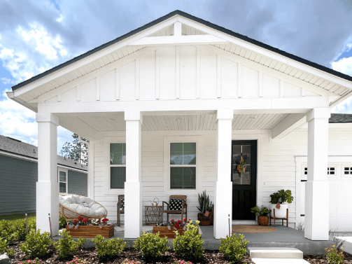 A white house with a covered front porch, potted plants, a papasan chair, a wicker chair, and a black front door stands under a partly cloudy sky in the inviting Wildlight Yulee Florida community.