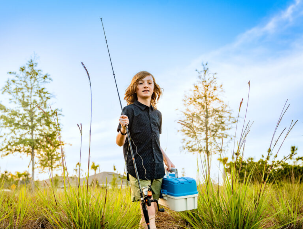 A boy with shoulder-length hair holds a fishing rod and tackle box, walking through tall grass outdoors under a blue sky in the scenic Wildlight Yulee Florida community.
