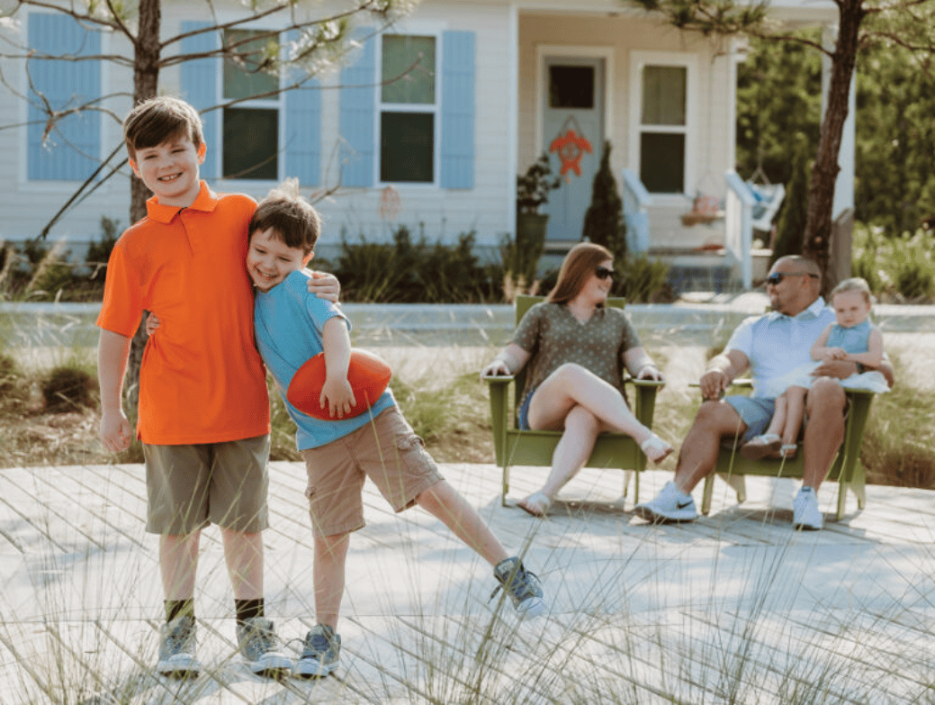 Two boys stand on a boardwalk, one hugging the other, while two adults and a child sit on chairs in front of a house—a glimpse into the warm spirit of the Wildlight Yulee Florida community and its inviting new homes in Wildlight FL.