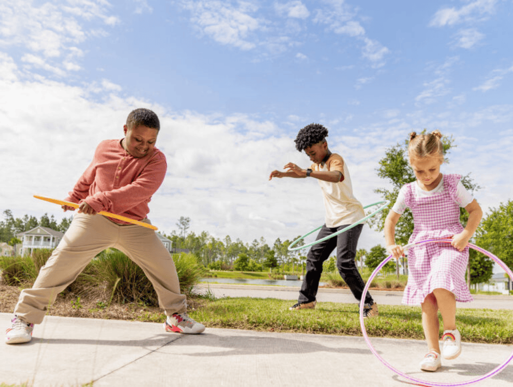 Three children hula hoop outdoors on a sunny day, enjoying the grassy areas and sidewalks of the Wildlight Yulee Florida community, with charming homes in Nassau County Florida and tree-lined streets in the background.