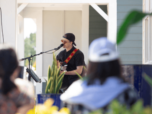 A musician wearing sunglasses and a backward cap plays guitar and sings into a microphone on an outdoor stage at the Wildlight Yulee Florida community, with audience members in the foreground.
