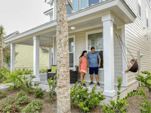 A couple stands on the porch of a house in the Wildlight master planned community while a child swings on a hanging chair nearby. The yard features plants and shrubs, reflecting the inviting lifestyle found in homes in Nassau County Florida.