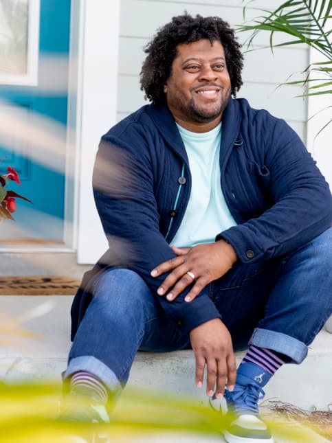 Man with curly hair, wearing a navy cardigan, light blue shirt, and jeans, sits on a porch step of one of the new homes in Wildlight FL, smiling with plants visible in the foreground and background.