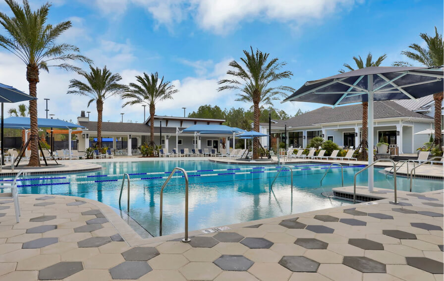 Outdoor swimming pool area with lounge chairs, umbrellas, and palm trees, surrounded by adjacent buildings under a partly cloudy sky in the vibrant Wildlight Yulee Florida community.