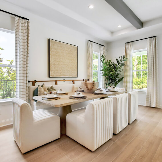 Modern dining area with a wooden table, six white upholstered chairs, a built-in bench, neutral wall art, large windows with sheer curtains, and a tall potted plant—perfect for new homes in Wildlight FL.