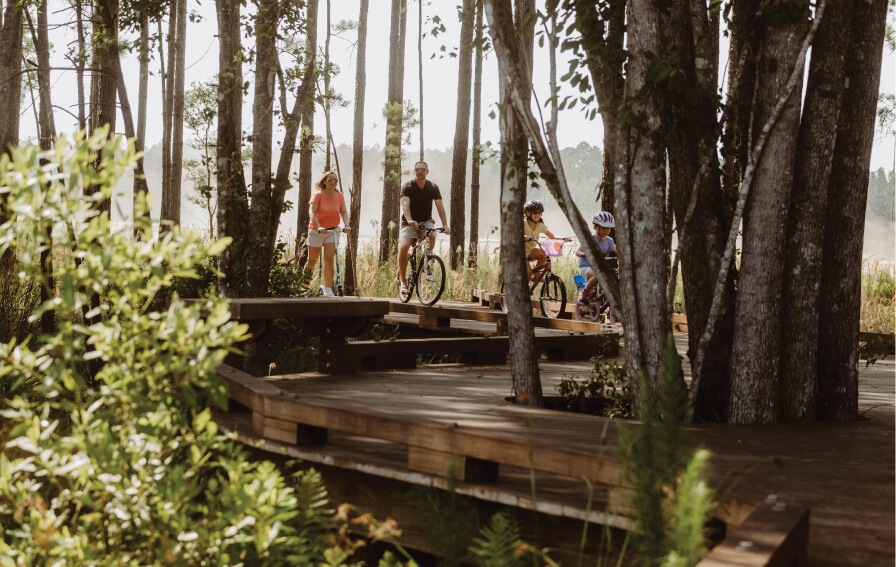 Four people, including two children on bikes and two adults walking, travel along a wooden boardwalk winding through the Wildlight Yulee Florida community, surrounded by trees and greenery.
