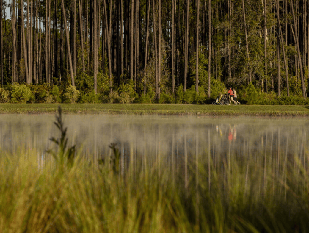 A person with a bicycle stands on a grassy path beside a calm, reflective body of water in the scenic Wildlight master planned community, with tall trees providing a tranquil backdrop.