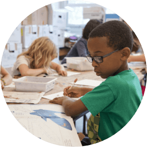 Several children sit at a classroom table, focused on writing and drawing activities in Wildlight Florida. One boy in a green shirt and glasses works in the foreground, highlighting the vibrant community life found near new homes in Wildlight FL.