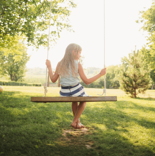 A young girl with long blonde hair sits barefoot on a wooden swing in a sunlit, grassy yard surrounded by trees in the welcoming Wildlight Yulee Florida community.