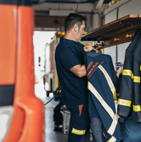 A firefighter in uniform stands in a garage, holding a fire jacket with reflective stripes, ready to serve the Wildlight Yulee Florida community. Firefighting equipment and vehicles are visible in the background.