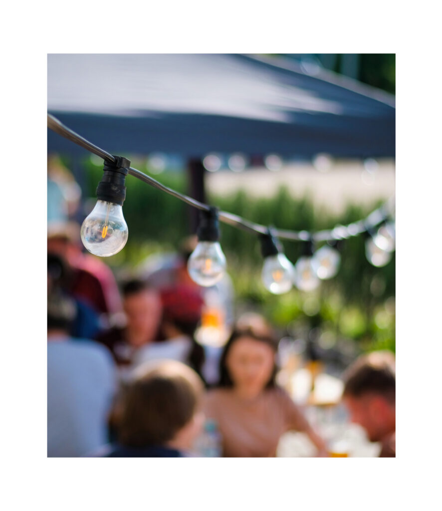 A string of clear light bulbs hangs outdoors above a group of people sitting and talking under a canopy on a sunny day in the vibrant Wildlight master planned community.