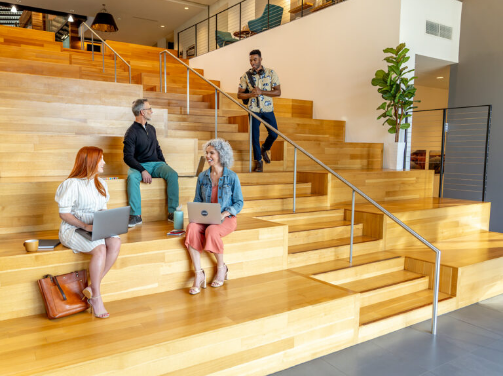 Four people are sitting and standing on wide wooden steps in a modern office at Wildlight Yulee Florida community, some using laptops and conversing about new homes in Wildlight FL.