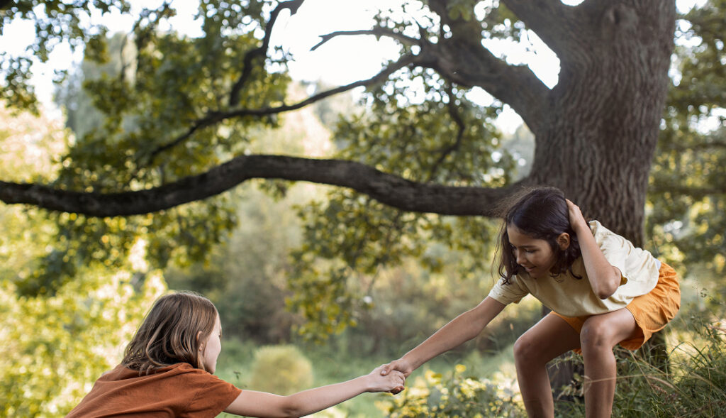 Two children outdoors, one helping the other up a grassy hill near a large tree with green foliage—a heartwarming scene that captures the spirit of camaraderie found in homes in Nassau County, Florida.