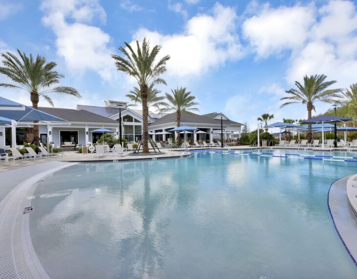 Outdoor swimming pool with clear water, surrounded by lounge chairs, palm trees, and umbrellas, in front of a modern clubhouse under a blue sky—perfect for relaxing in the vibrant Wildlight Yulee Florida community.
