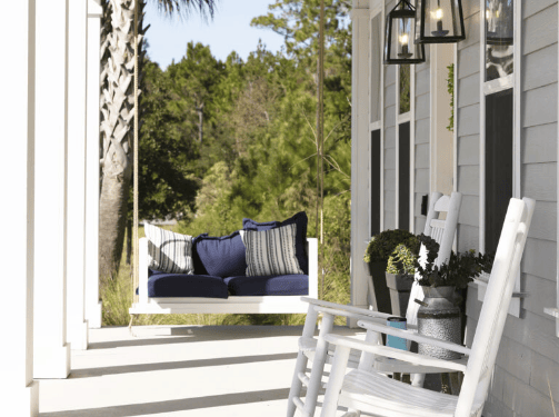 Charming white porch with a blue cushioned swing, white rocking chair, potted plants, and lantern-style lights—perfect for relaxing in the scenic Wildlight master planned community in Nassau County, Florida.