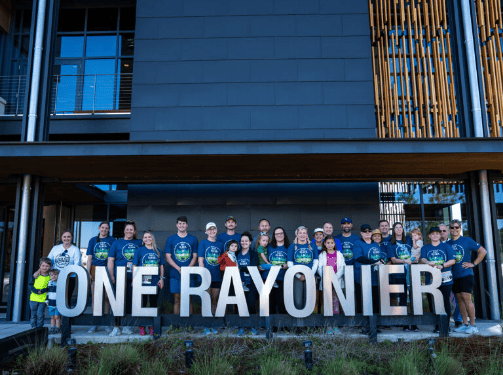 A group of people in matching shirts stand behind a large sign reading "ONE RAYONIER" in front of a modern building, celebrating the vibrant Wildlight master planned community and its new homes in Wildlight FL.