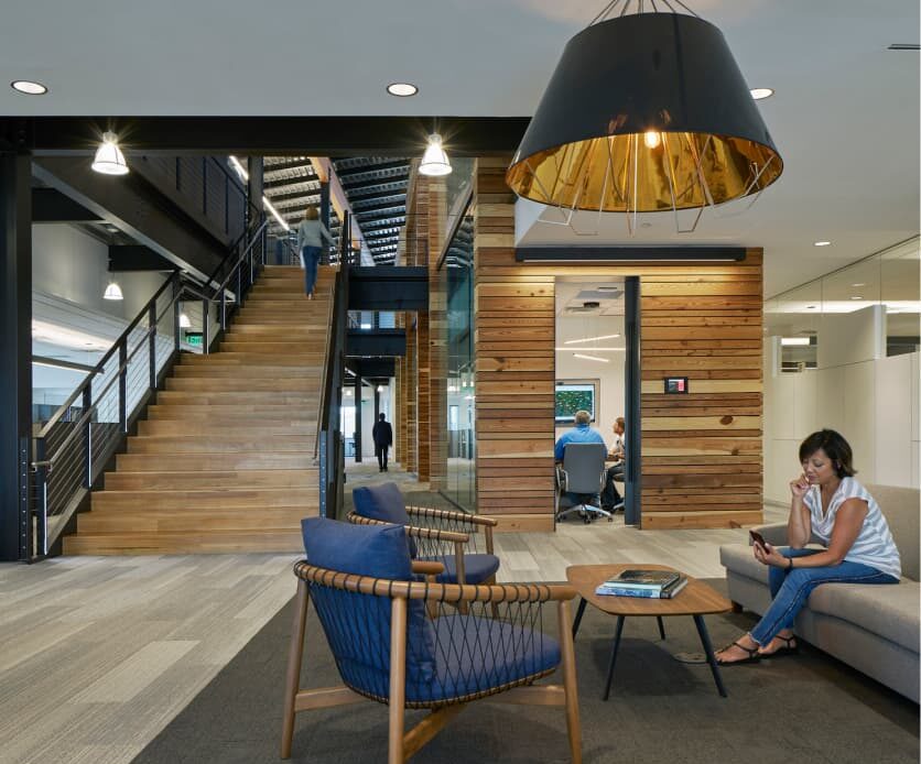 Modern office interior in the heart of the Wildlight master planned community, featuring a wooden staircase, lounge chairs, and a woman using a laptop. Glass-walled meeting room visible in the background highlights homes in Nassau County Florida.