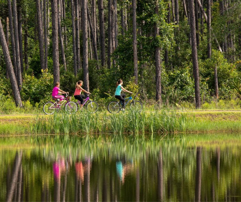 Three people ride bicycles on a path through a forested area beside a pond in the Wildlight Yulee Florida community, with their reflections visible in the water.