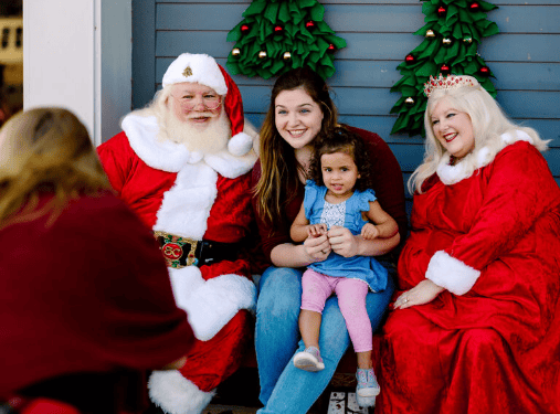A man dressed as Santa Claus, a woman as Mrs. Claus, a young woman, and a small girl sit together and smile in front of a decorated wall at Wildlight Florida, a vibrant master planned community in Nassau County.