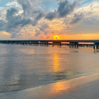 A wooden pier stretches over calm water at sunset in Wildlight Florida, with clouds above and the sun reflecting on the wet sand and sea.