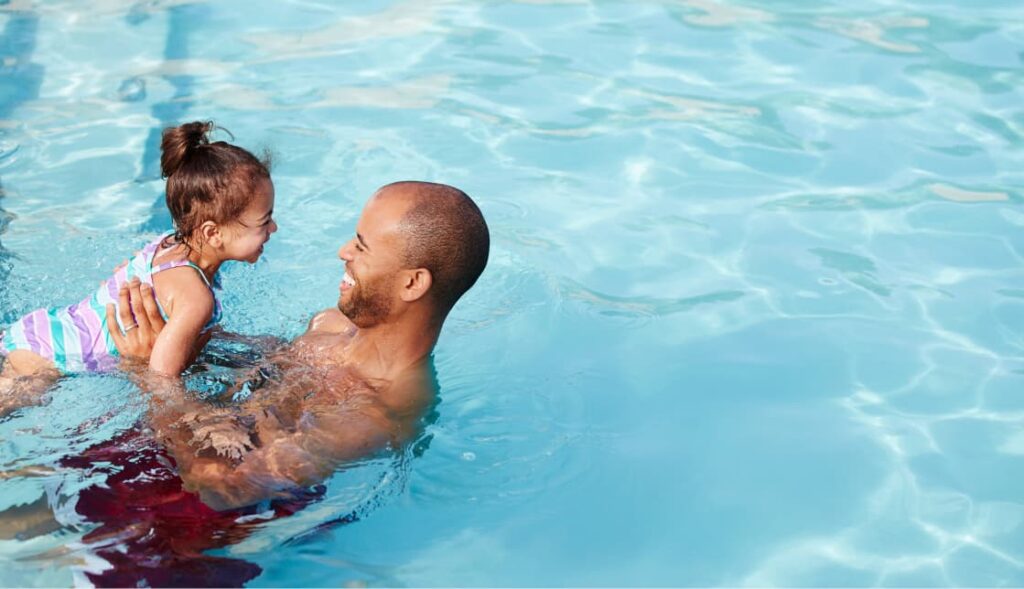 An adult holds a young child in a swimsuit while standing in a swimming pool filled with clear water, enjoying quality time at the Wildlight master planned community.