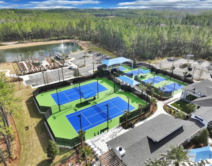 Aerial view of a sports complex with tennis and pickleball courts, surrounded by trees, a pond, parking lot, and nearby buildings in the vibrant Wildlight Yulee Florida community.