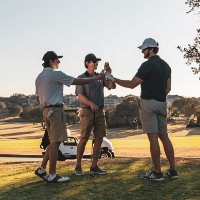 Three men standing on a golf course in the Wildlight Yulee Florida community clink bottles together, with a golf cart and trees visible in the background.