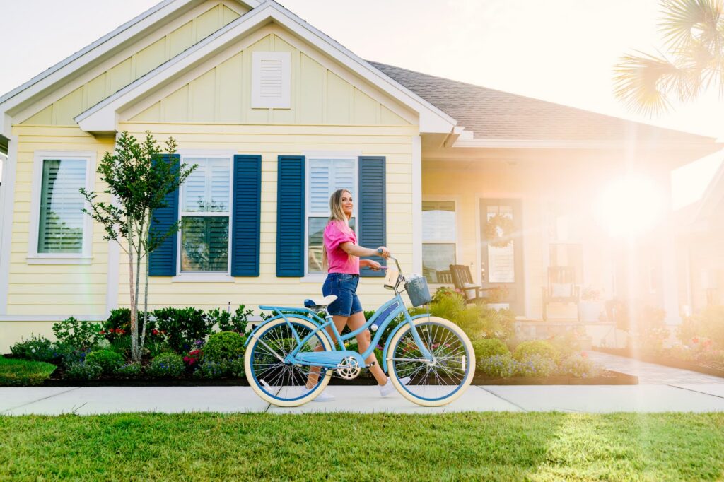 A woman walks a blue bicycle on the sidewalk in front of a yellow house with blue shutters on a sunny day, capturing the vibrant lifestyle found in new homes in Wildlight FL.