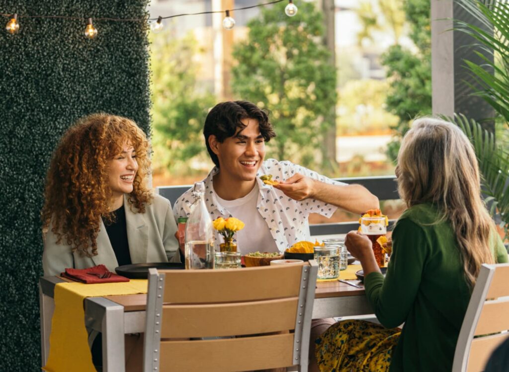 Three people sit at an outdoor table with food and drinks, talking and smiling amid greenery and string lights—capturing the relaxed lifestyle of the Wildlight master planned community in Wildlight, Florida.