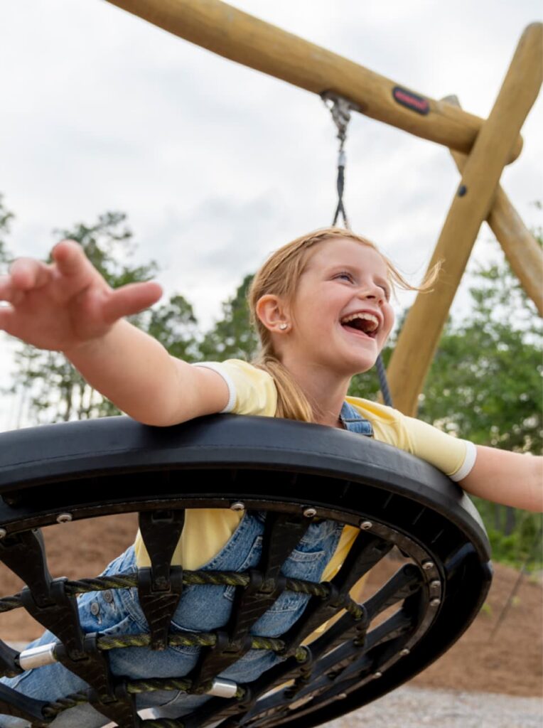 A young girl with blond hair, wearing a yellow shirt and overalls, smiles and stretches her arms while swinging on a round swing at an outdoor playground in Wildlight Florida.