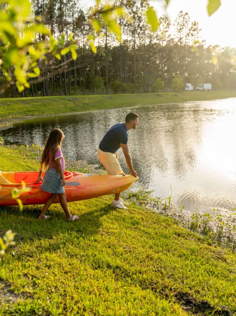 A man and a young girl stand by a lake in the Wildlight Yulee Florida community, preparing to launch a yellow kayak onto the water on a sunny day.