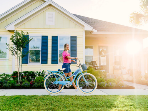 A woman in a pink shirt and shorts walks a blue bicycle on a sidewalk in front of a yellow house within the vibrant Wildlight Yulee Florida community on a sunny day.