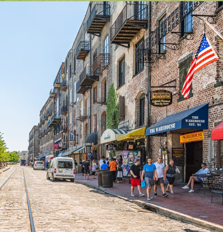 People walk along a cobblestone street lined with historic brick buildings, shops, and an American flag in charming Wildlight Florida. Some cars are parked on the side of the road near old rail tracks, blending history with new homes in Wildlight FL.