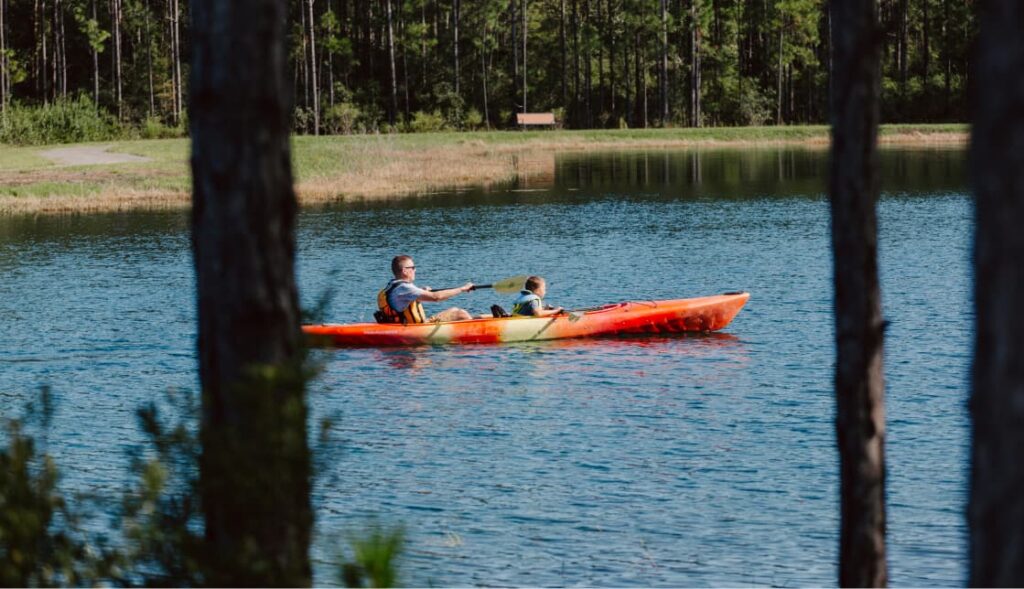 Two people are kayaking on a calm lake surrounded by trees in the Wildlight Yulee Florida community, with a grassy area and bench visible in the background.
