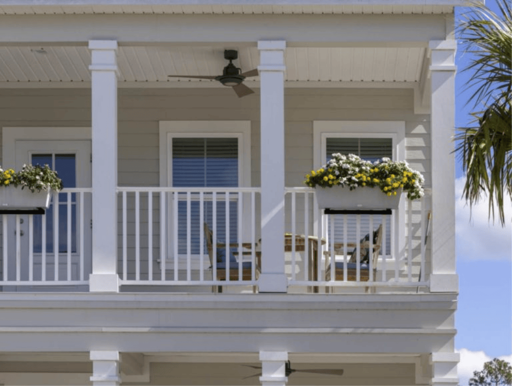 Second-story porch with white railing, two windows, a ceiling fan, a table with two chairs, and flower boxes brimming with yellow and white flowers in the inviting Wildlight Yulee Florida community.