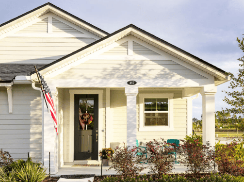 Charming single-story white house with a front porch, black door, potted plants, two blue chairs, and an American flag—perfect for those seeking new homes in Wildlight FL.