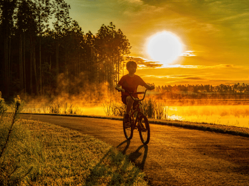 A child rides a bicycle on a paved path beside a lake at sunrise, with trees and mist in the background, reflecting the serene lifestyle found in the Wildlight Yulee Florida community.
