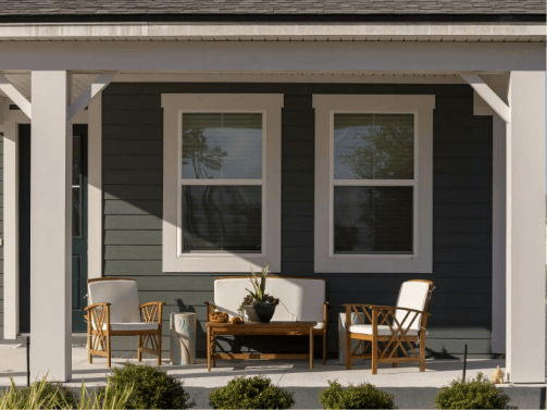 A front porch in the Wildlight Yulee Florida community features two windows, a wooden outdoor seating set, a coffee table with a potted plant, and green shrubs in the foreground.