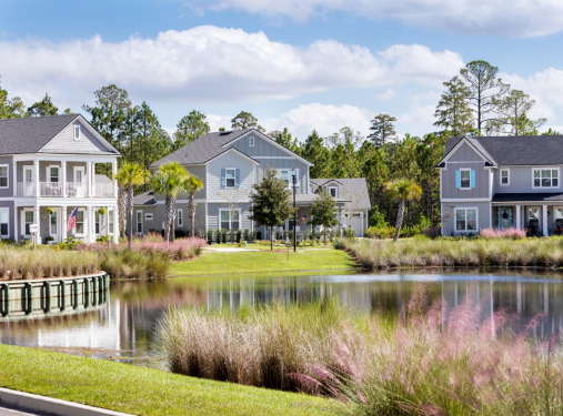Modern suburban houses with front porches overlook a pond and landscaped grass, set among trees under a partly cloudy sky—perfect examples of new homes in Wildlight FL.