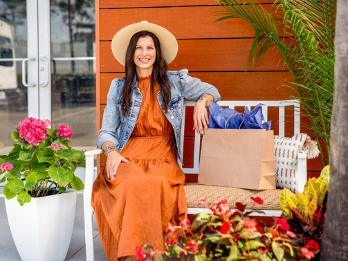 Woman in a hat and orange dress sits on a bench with a shopping bag, surrounded by flowers and plants, in front of a wooden wall—capturing the relaxed vibe of the Wildlight master planned community.