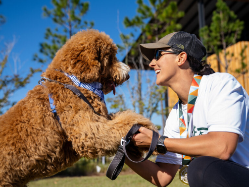A person wearing sunglasses kneels outdoors in Wildlight Florida, smiling and shaking hands with a large, curly-haired dog.