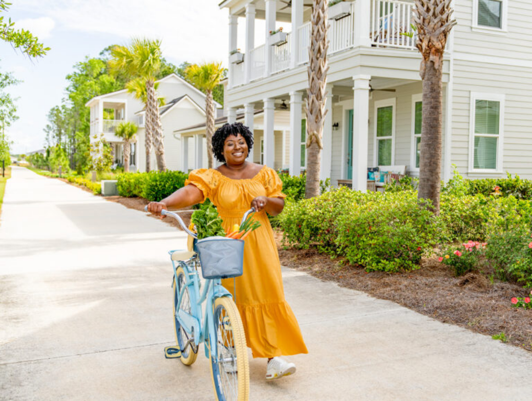 A woman in a yellow dress walks a blue bicycle with a basket of vegetables along a suburban sidewalk lined with homes in Nassau County Florida and palm trees, capturing the charm of the Wildlight Florida community.