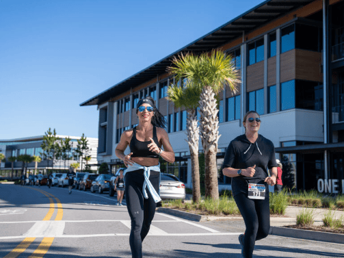 Two women in athletic wear run on a sunny street lined with modern buildings during a race event in the vibrant Wildlight Yulee Florida community.