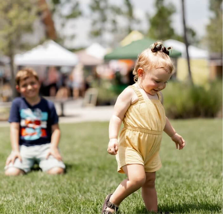 A young child in a yellow romper walks on grass while an older child sits in the background at an outdoor event with tents in the Wildlight Yulee Florida community.