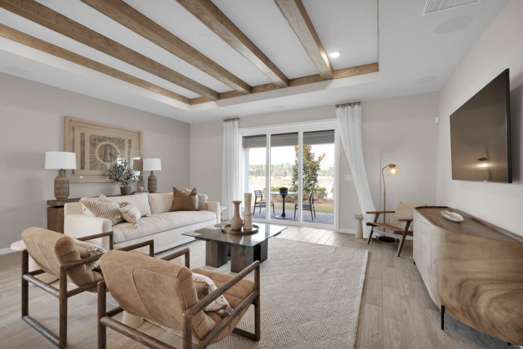 A modern living room in the Wildlight Yulee Florida community features wooden ceiling beams, neutral furnishings, a wall-mounted TV, glass coffee table, and sliding doors opening to a patio.