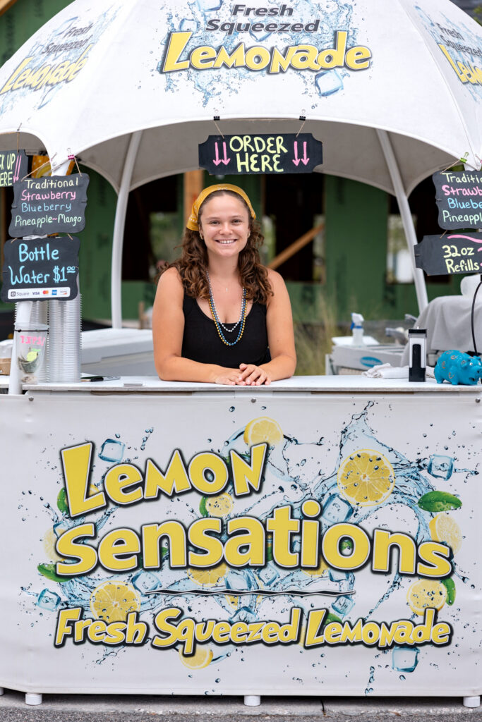 A woman stands at a lemonade stand called "Lemon Sensations," offering fresh squeezed lemonade and bottled water under a white canopy in the Wildlight Yulee Florida community.