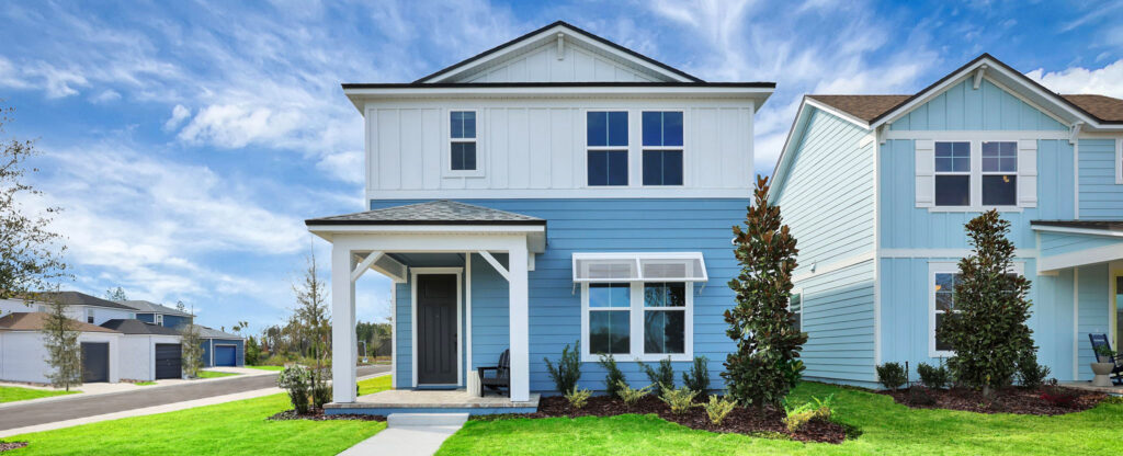 A two-story blue and white house with a covered front porch, manicured lawn, and neighboring homes in the Wildlight master planned community under a partly cloudy sky.