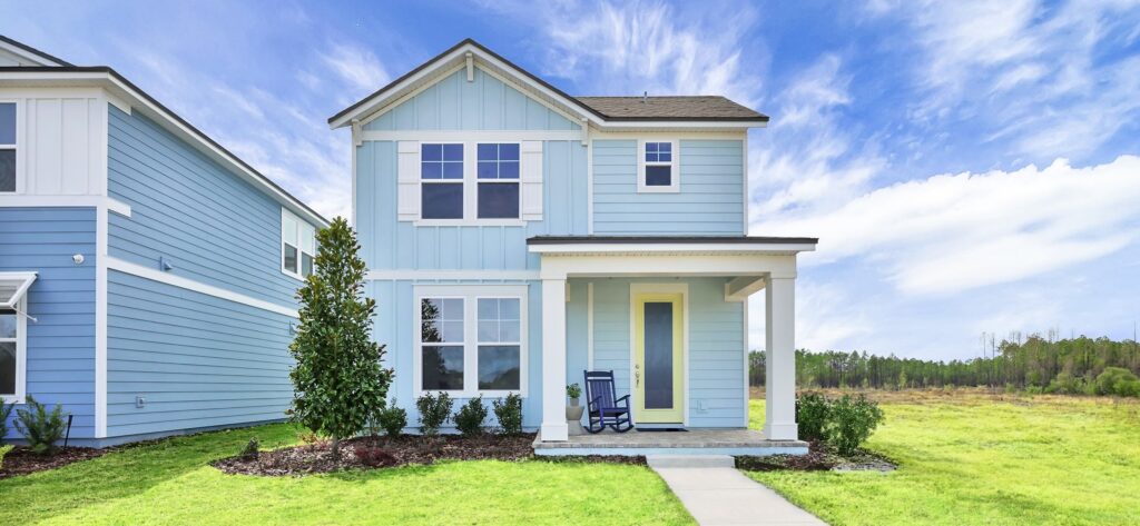 A light blue two-story house with white trim, a front porch, rocking chair, and manicured landscaping sits in the Wildlight Yulee Florida community, surrounded by grassy open spaces and blue sky.